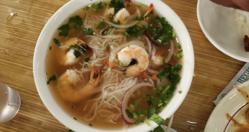 A bowl of shrimp noodle soup with herbs and vegetables, served on a wooden table.