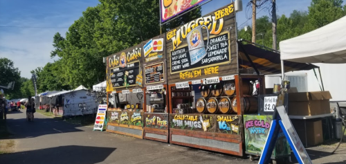 A colorful food stand at a fair, featuring signs for lemonade, snacks, and drinks, with people walking by.