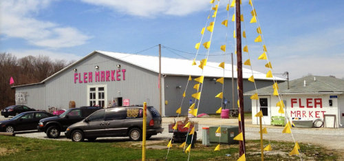 A large gray building with "Flea Market" signage, surrounded by vehicles and colorful flags on a sunny day.