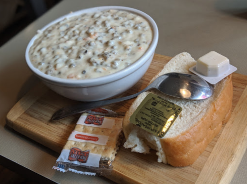 A bowl of creamy soup with meat and vegetables, served with a slice of bread and butter on a wooden cutting board.