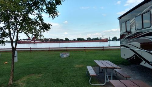 A riverside scene featuring an RV, picnic table, and a large ship passing by on the water. Green grass and trees are visible.