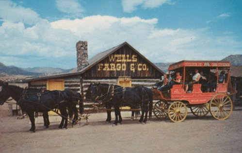 A vintage stagecoach pulled by horses in front of a Wells Fargo building, with passengers enjoying the ride.