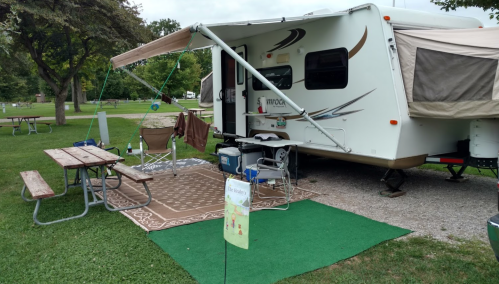 A cozy campsite featuring a camper, picnic table, chairs, and a green mat on a grassy area.