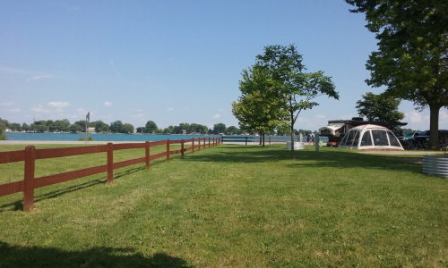 A grassy area by a lake, featuring a wooden fence, a tree, and a camping tent in the background under a clear blue sky.