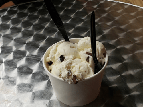 A cup of vanilla ice cream with chocolate chunks, served with two black plastic spoons on a metallic table.