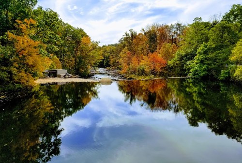 A serene river scene reflecting colorful autumn trees under a blue sky with scattered clouds.