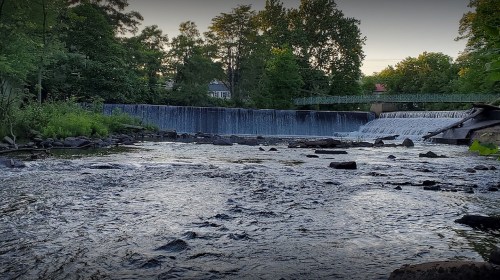 A serene river scene with a waterfall, surrounded by lush greenery and a bridge in the background.