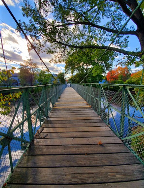 A wooden pedestrian bridge surrounded by trees and water, under a blue sky with scattered clouds.