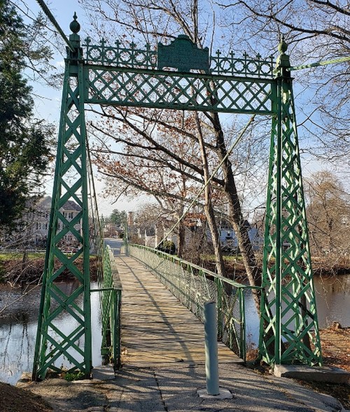 A green, ornate suspension bridge spans a calm river, surrounded by trees and a clear blue sky.