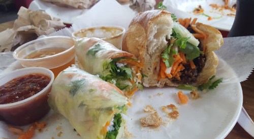 A plate with a partially eaten sandwich and fresh spring rolls, accompanied by dipping sauces.