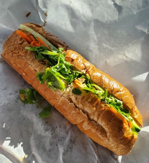 A fresh sandwich on a paper wrapper, filled with vegetables like carrots, cucumber, and cilantro on whole grain bread.