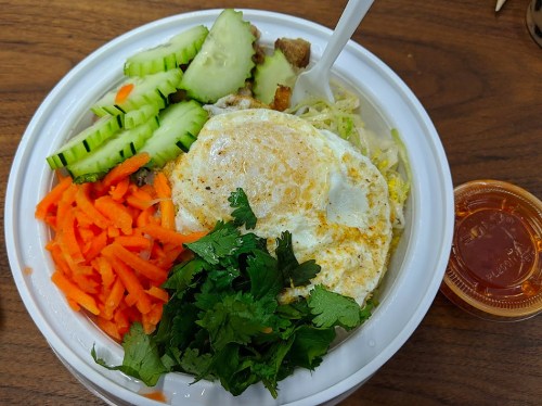 A bowl of rice topped with a fried egg, carrots, cucumbers, lettuce, and cilantro, served with a side of sauce.