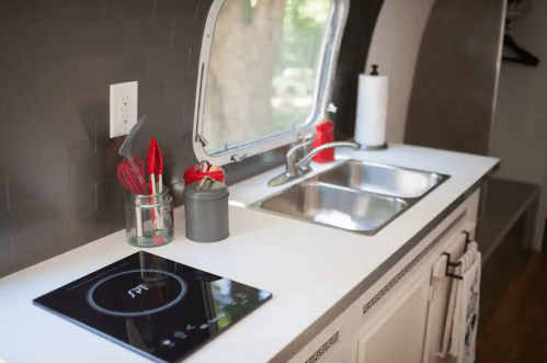 A modern kitchen with a sink, stovetop, and utensils in jars, featuring a large window with a view of trees outside.