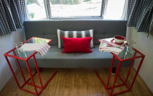 Cozy seating area with a gray couch, red pillows, and two red side tables with glass tops, set in a bright room.