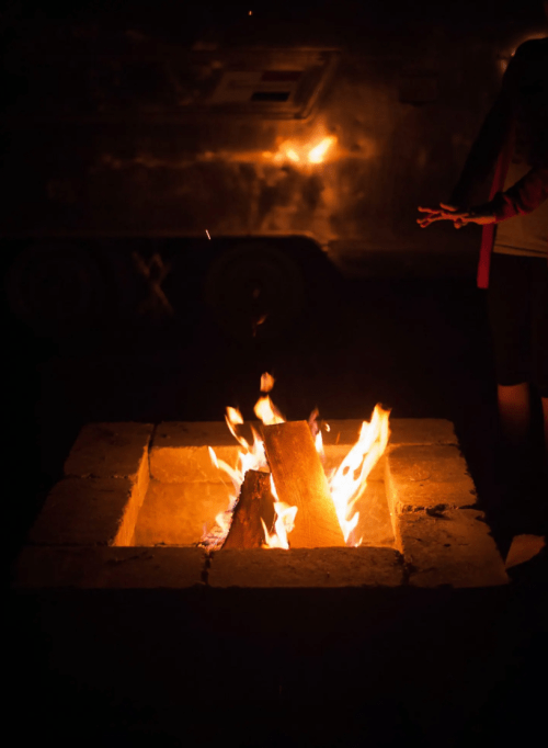 A cozy campfire burns in a stone fire pit, with a person’s hand reaching out nearby in the dark.