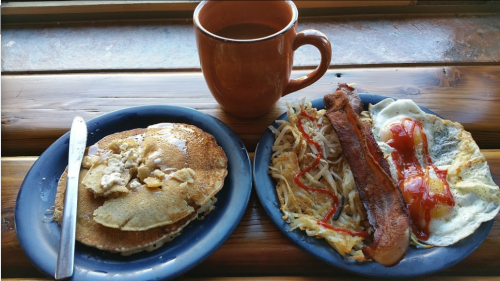 A plate with pancakes, a side of eggs, hash browns, and a sausage, accompanied by a cup of coffee.