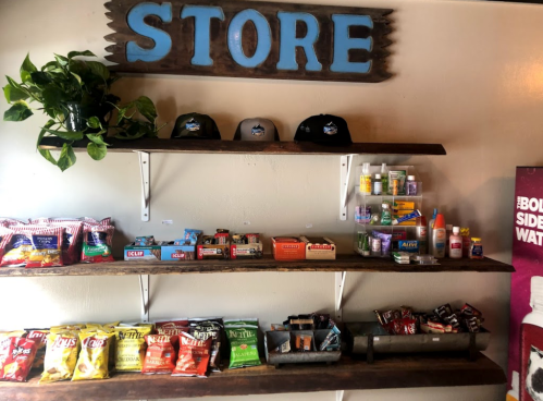 A rustic store display with shelves of snacks, drinks, and hats, featuring a plant and a sign that reads "STORE."