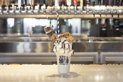 A tall milkshake topped with whipped cream, chocolate drizzle, a cookie, and two straws, set against a bar backdrop.