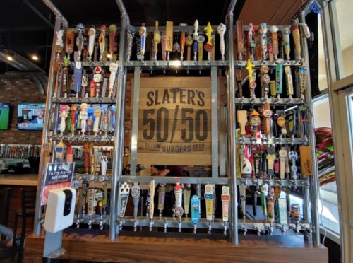 A wall display of various beer taps at Slater's 50/50, featuring a sign that reads "Slater's 50/50 Bacon Burgers Beer."