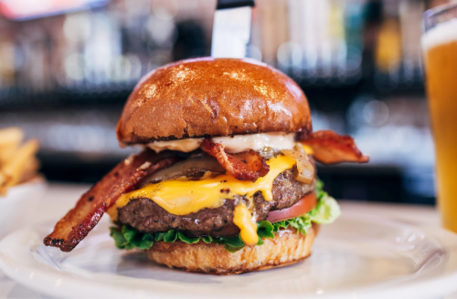 A juicy cheeseburger with bacon, lettuce, tomato, and a glossy bun, served on a plate with a beer in the background.