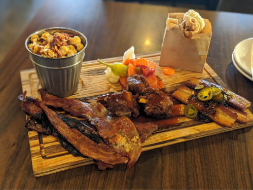 A wooden platter featuring crispy bacon, meatballs, pickled vegetables, and a side of popcorn in a cup.