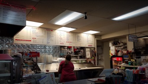 A woman stands at a counter in a cozy diner, with a menu board and various kitchen items visible in the background.