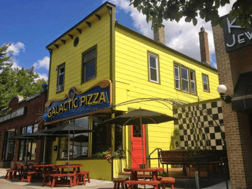 Bright yellow building with "Galactic Pizza" sign, outdoor seating, and checkered wall, set against a blue sky.