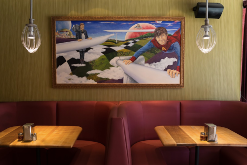 A colorful mural featuring two figures on a surreal landscape, above cozy red booths with wooden tables in a restaurant.