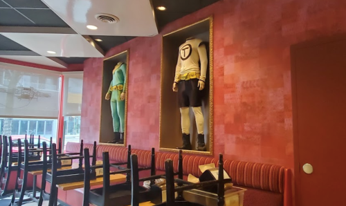 Two superhero costumes displayed in frames on a red wall inside a restaurant, with chairs stacked upside down on tables.