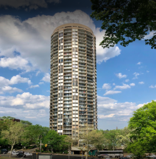 A tall, cylindrical residential building surrounded by trees under a partly cloudy sky.