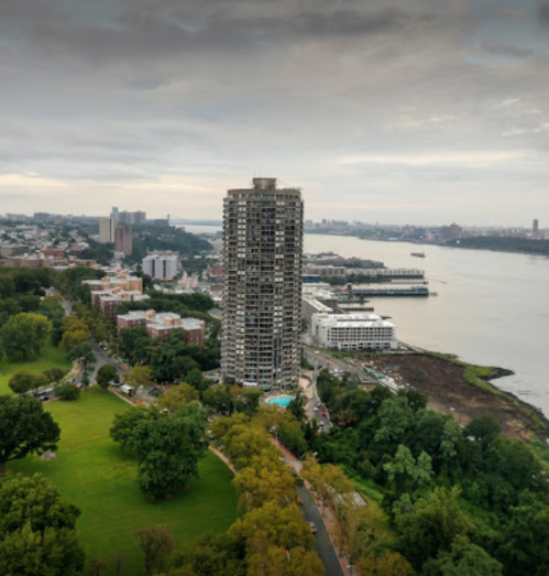 Aerial view of a tall building near a river, surrounded by green parks and cityscape under a cloudy sky.