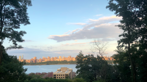 A scenic view of a city skyline at sunset, framed by trees and reflecting on a calm body of water.