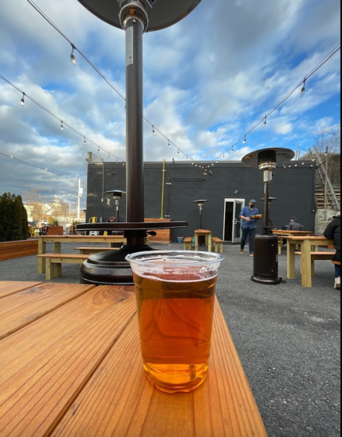 A clear cup of beer sits on a wooden table in an outdoor seating area with string lights and heaters.