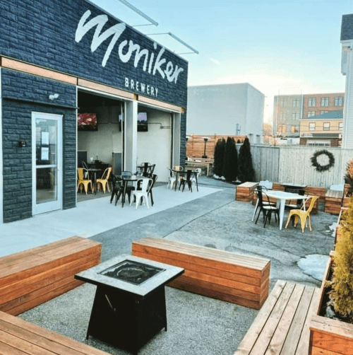Outdoor seating area at Moniker Brewery, featuring wooden benches, a fire pit, and a modern building facade.