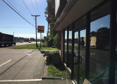 A view of a roadside building with a sign, parking area, and a truck passing by on a sunny day.