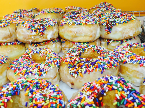 A close-up of a pile of glazed donuts topped with colorful sprinkles against a bright orange background.