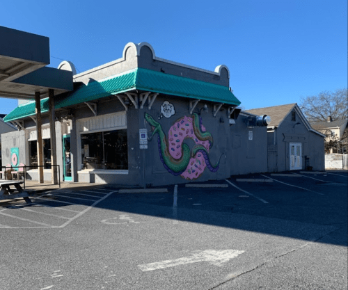 A colorful mural of a snake wrapped around a donut on the side of a building with a teal roof and empty parking lot.
