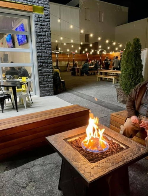 A cozy outdoor space with a fire pit, string lights, and people dining in the background.