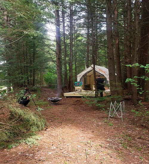 A tent set up in a forest, with a person nearby and trees surrounding the area. Sunlight filters through the branches.