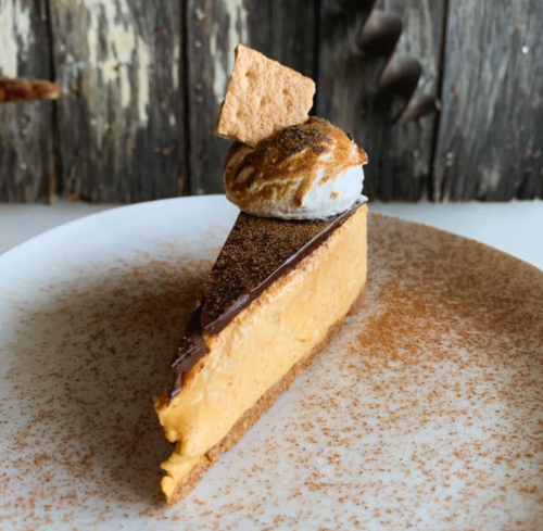 A slice of creamy dessert topped with a toasted meringue and a graham cracker on a textured plate.