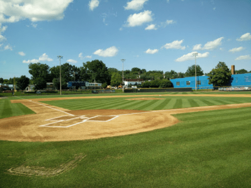 A well-maintained baseball field with a clear blue sky and fluffy clouds in the background.