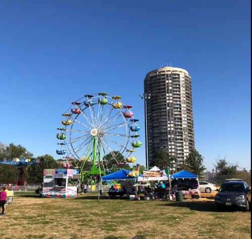 A colorful ferris wheel at a fair, with food stalls and a tall building in the background under a clear blue sky.