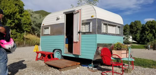 A vintage teal and white caravan parked on gravel, with red chairs and a small wooden deck outside.