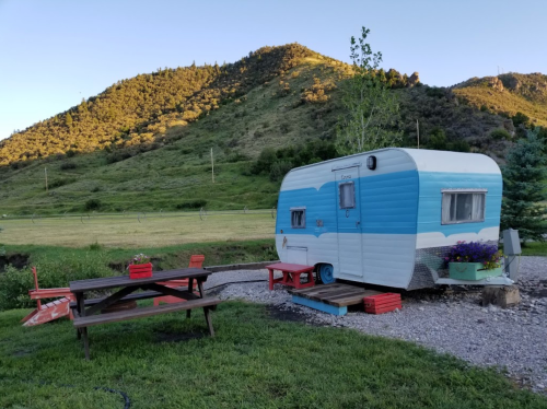 A vintage blue and white camper trailer sits beside a picnic table in a grassy area with mountains in the background.