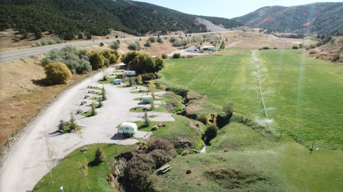 Aerial view of a campground with RVs, green fields, and mountains in the background on a sunny day.