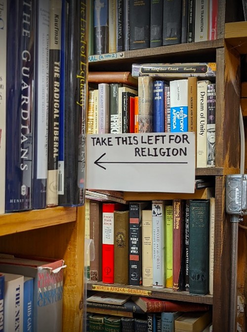 A sign on a bookshelf directing to the left for books on religion, surrounded by various other books.