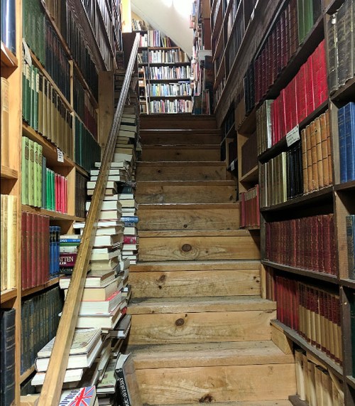 A wooden staircase surrounded by tall bookshelves filled with colorful books, with stacks of books on the steps.