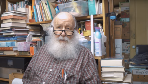An elderly man with a long beard and glasses sits in a cluttered room filled with books and various items.