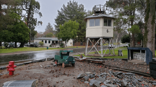 A small green toy truck near a weathered watchtower, surrounded by trees and a rainy street scene.