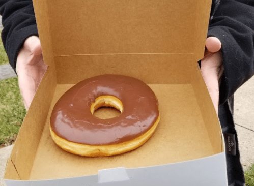 A person holding a box with a single chocolate-glazed donut inside.